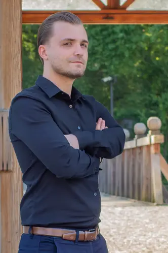 Wyprostowany mężczyzna dumnie pozuje na tle parku. Ubrany jest w ciemną koszulę włożoną w spodnie.